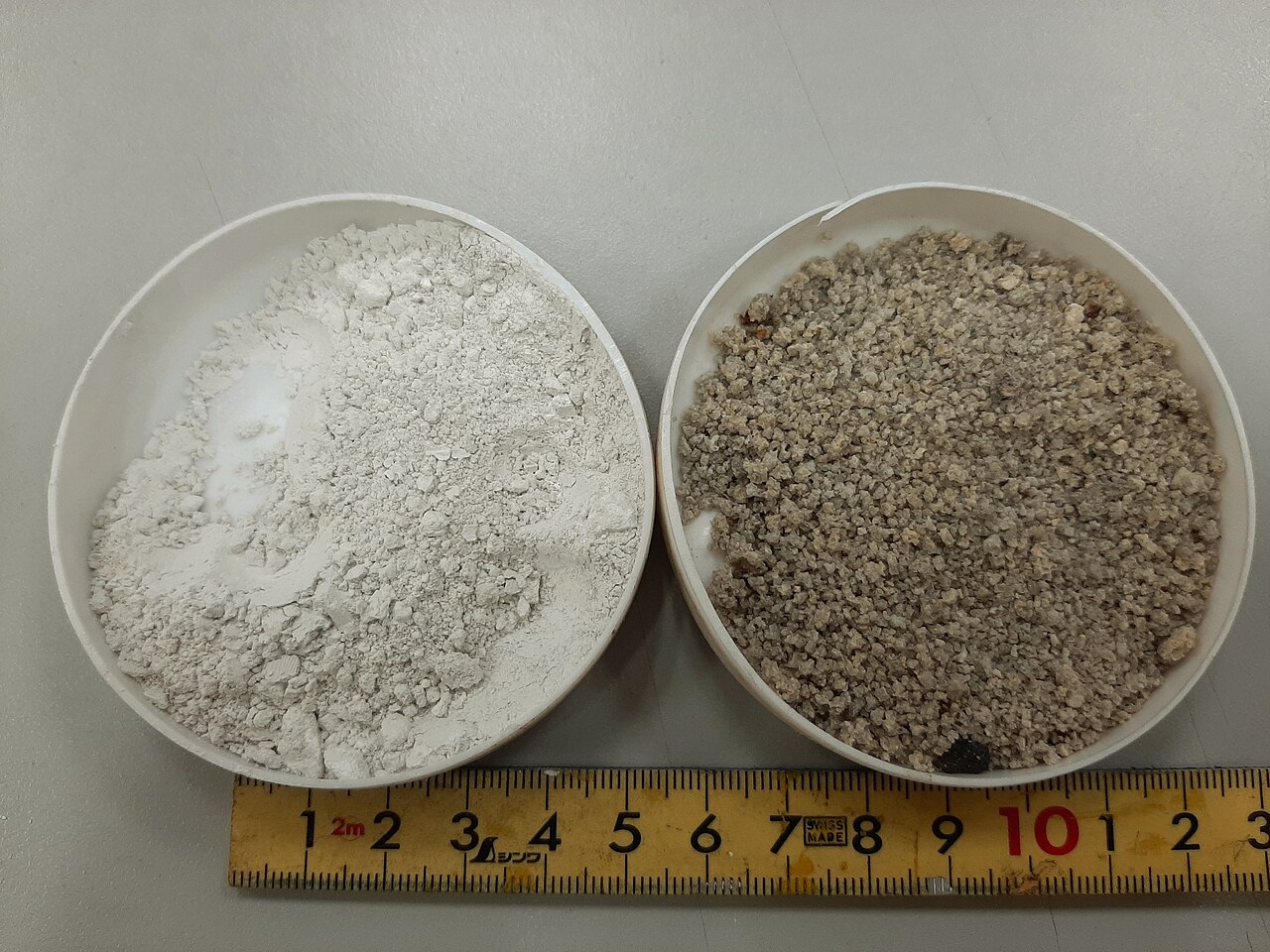 Two dishes of blast-furnace slag: a pale ground powder and coarser granulated slag beside a ruler