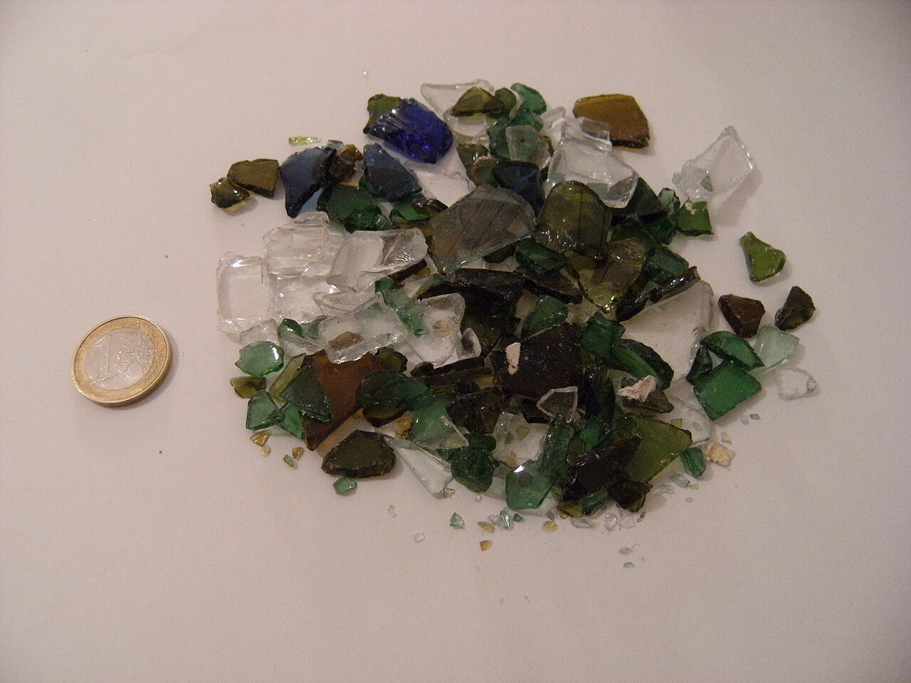 Mixed-colour crushed container-glass cullet on a white surface beside a euro coin