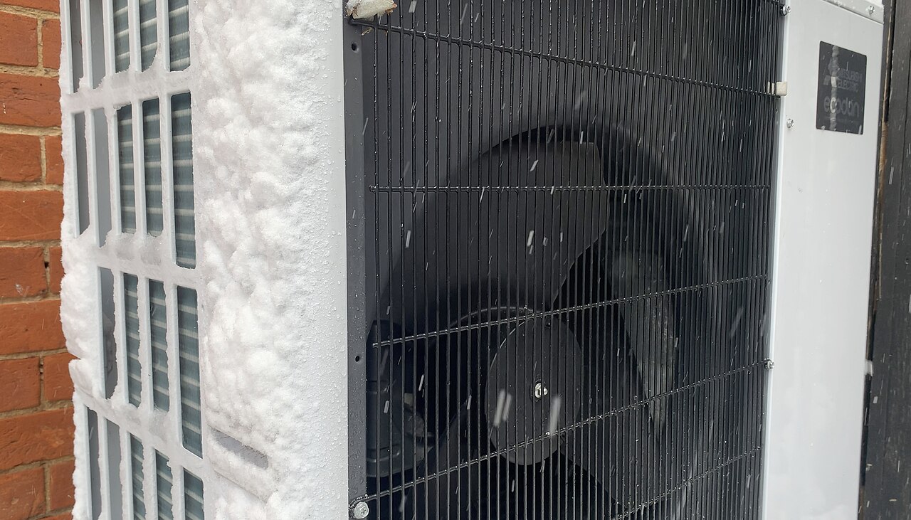 An outdoor air-source heat pump unit running beside a brick wall in falling snow