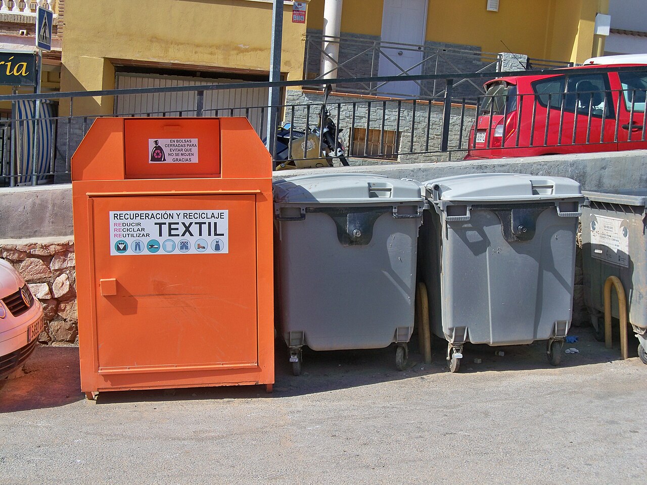 An orange street container labeled for textile recovery and recycling beside grey waste bins